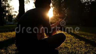 公园日落时分，一个手里拿着智能<strong>手机</strong>的人坐在草地上的<strong>剪影</strong>